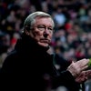 Sir Alex Ferguson watching a football game from the side of the pitch while a crowd of fans watch in the background