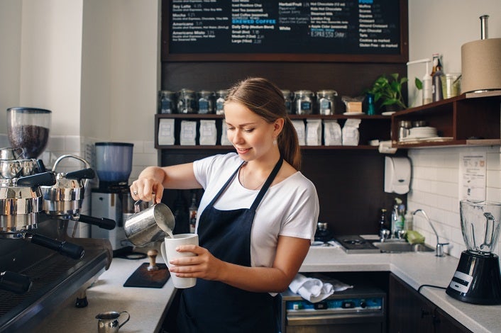 Portrait of smiling young Caucasian woman pouring hot milk in coffee. Waitress holding white mug cup in cafe. Person at work, small business concept